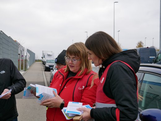 Zwei Frauen in roten Warnwesten stehen auf einer Straße und beugen sich gemeinsam über Infromationsmaterialien. 